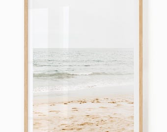 May include: A framed print depicting a tranquil beach vista. The photograph shows the sea meeting the sandy beach under a muted, overcast sky. The frame is a light wood colour, providing a natural aesthetic to the coastal artwork.