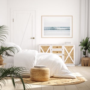 May include: A white bedroom with a bed, a dresser, and a rug. The bed has white linens and a white throw pillow. The dresser has a white top and a woven rattan front. The rug is woven and has a natural color. There is a framed photograph of a beach scene above the dresser. There are two potted plants in the room, one on the floor and one on a table.