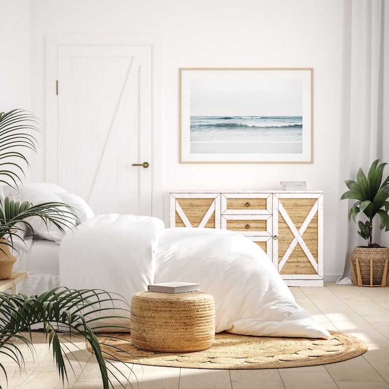 May include: A white bedroom with a bed, a dresser, and a rug. The bed has white linens and a white throw pillow. The dresser has a white top and a woven rattan front. The rug is woven and has a natural color. There is a framed photograph of a beach scene above the dresser. There are two potted plants in the room, one on the floor and one on a table.