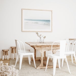 May include: A white dining table with four white metal chairs and a framed photograph of a beach scene above it. The table is made of wood and has a rustic finish. The chairs have a simple design and are comfortable. The photograph is a beautiful depiction of a beach with blue water and white sand.