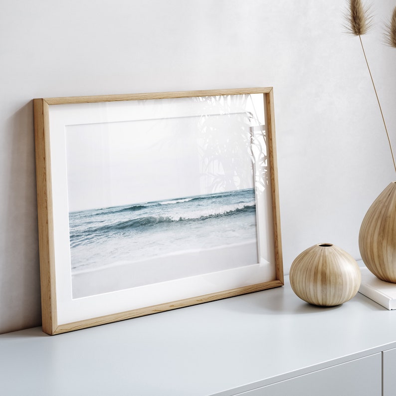 May include: A framed photograph of a beach scene with blue waves crashing on the shore. The frame is made of light wood and has a white mat.