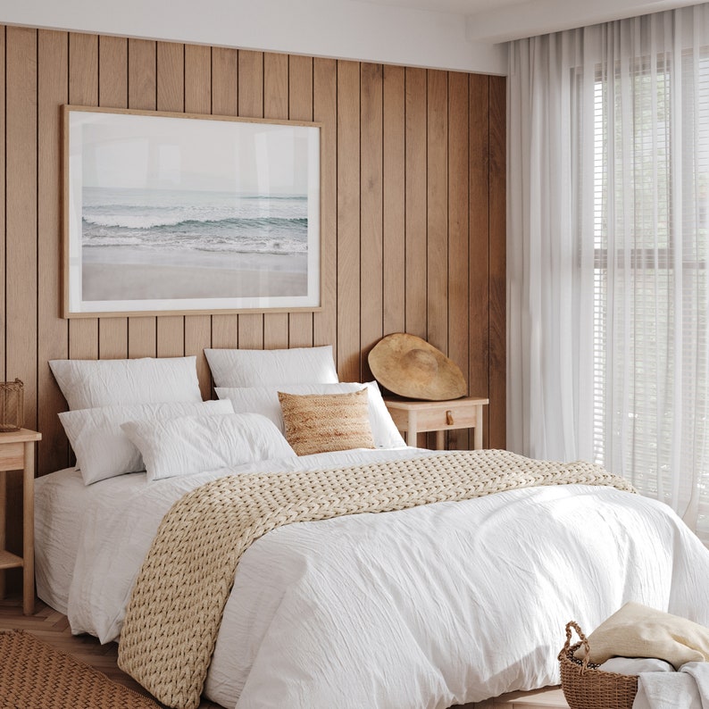 May include: A white bed with a white duvet cover and a beige knitted throw blanket. The bed has two white pillows and a beige pillow with a woven texture. The bed is in a bedroom with wood paneling on the walls and white curtains.