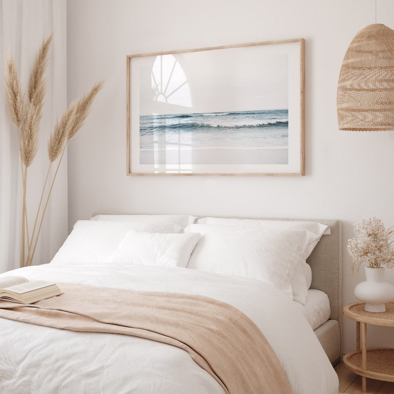 May include: A white bed with a tan blanket and white pillows. A framed photograph of a beach scene hangs above the bed. A wicker lampshade hangs from the ceiling.