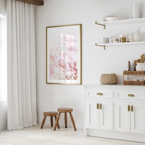 Pu&ograve; includere: Una cucina bianca con una stampa di peonia rosa incorniciata in oro, due sgabelli in legno e un armadio bianco con ferramenta dorata.