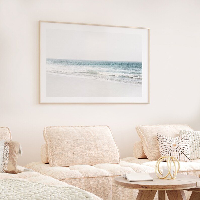 May include: A framed photograph of a beach scene with a light blue ocean and white sand. The photo is hanging on a white wall above a light beige sofa with two cushions.