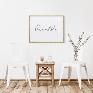 May include: A framed print with the word "breathe" in cursive script hangs above a small wooden table. Two white metal chairs flank the table. A vase of greenery and a candle sit on the table.