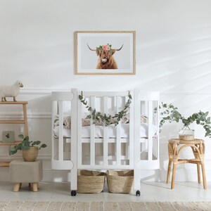 May include: White crib with a curved design and a white mattress. The crib has a garland of greenery hanging from the top. There is a small white sheep toy on a shelf to the left of the crib.