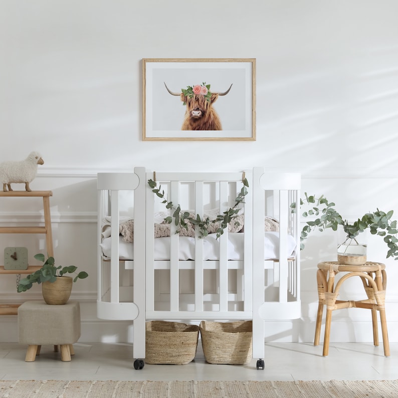 May include: White crib with a curved design and a white mattress. The crib has a garland of greenery hanging from the top. There is a small white sheep toy on a shelf to the left of the crib.
