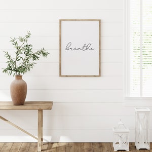 May include: A framed print with the word "breathe" in a cursive font hangs on a white shiplap wall. A wooden console table holds a terracotta vase with greenery. Two white lanterns sit on the wooden floor near a window.
