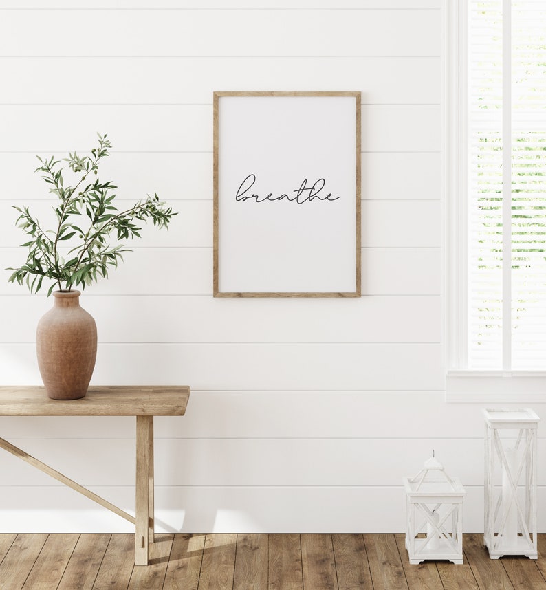 May include: A framed print with the word "breathe" in a cursive font hangs on a white shiplap wall. A wooden console table holds a terracotta vase with greenery. Two white lanterns sit on the wooden floor near a window.