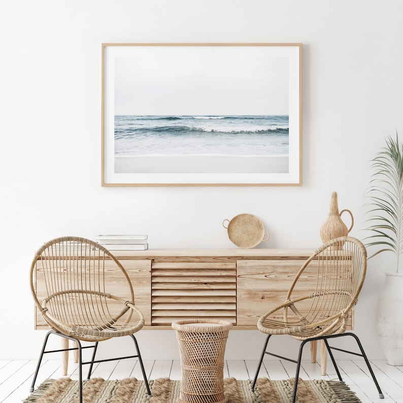 May include: A framed photograph of a beach scene with waves crashing on the shore. The photo is hanging on a white wall above a wooden cabinet with two wicker chairs and a small wicker table.