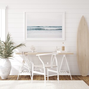 May include: A white desk with a white chair and a surfboard leaning against a white wall. The desk has a framed photo of a beach scene, a vase with a plant, and a few decorative items.