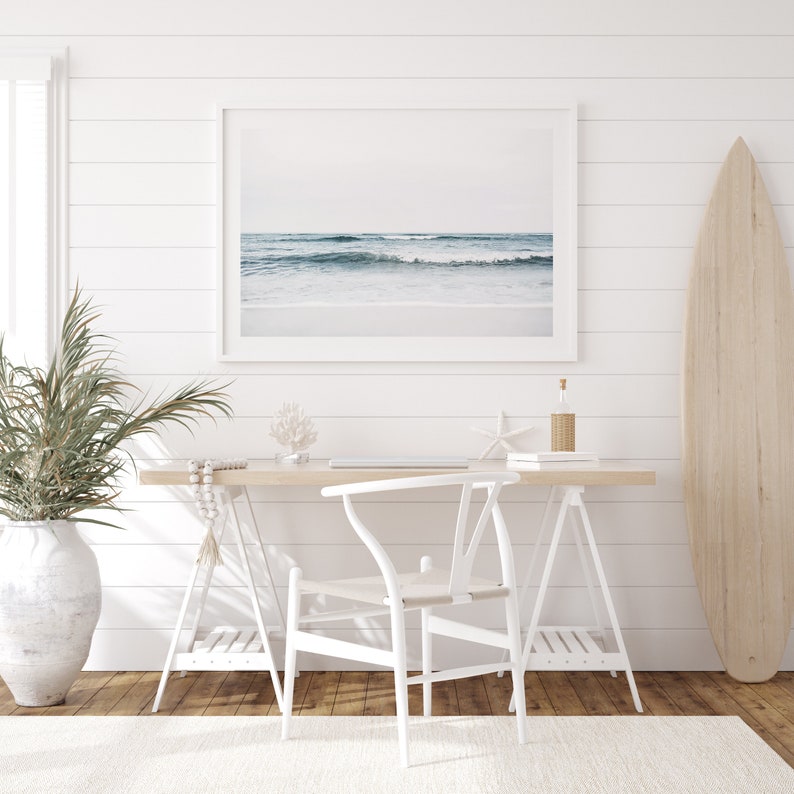 May include: A white desk with a white chair and a surfboard leaning against a white wall. The desk has a framed photo of a beach scene, a vase with a plant, and a few decorative items.