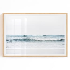 May include: A framed photograph of a seascape with blue waves crashing on a white sandy beach. The sky is a light blue and the horizon is visible in the distance.