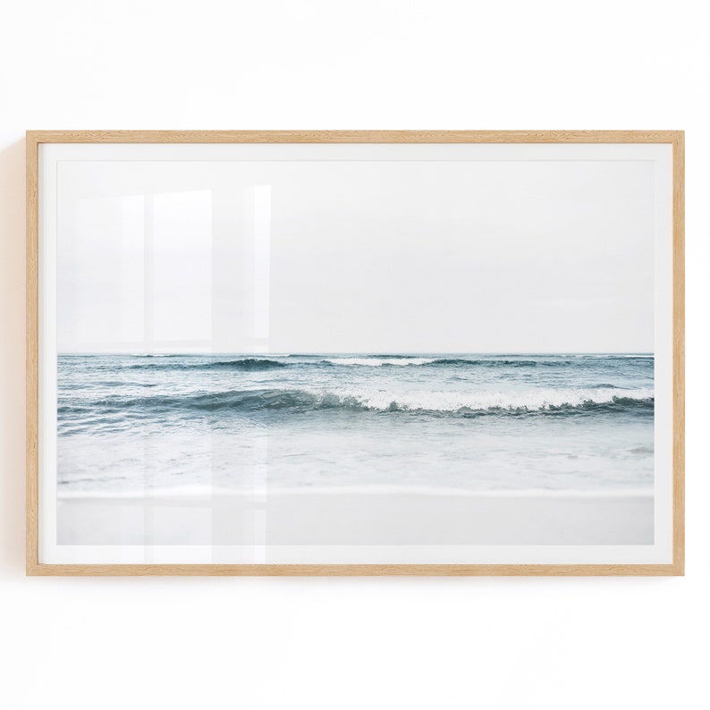 May include: A framed photograph of a seascape with blue waves crashing on a white sandy beach. The sky is a light blue and the horizon is visible in the distance.