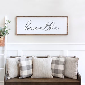 May include: A rectangular wooden sign with the word "breathe" in black script is mounted above a bench. Five decorative pillows in neutral tones, some with stripes and others with a plaid pattern, are arranged on the bench. A white ladder with a basket of greenery is on the left.