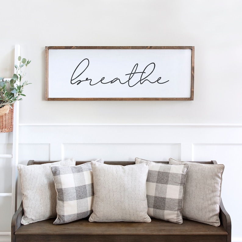 May include: A rectangular wooden sign with the word "breathe" in black script is mounted above a bench. Five decorative pillows in neutral tones, some with stripes and others with a plaid pattern, are arranged on the bench. A white ladder with a basket of greenery is on the left.