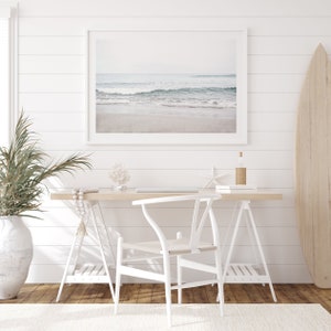 May include: A white desk with a white chair and a framed photograph of a beach scene. There is a surfboard leaning against the wall and a potted plant on the floor.