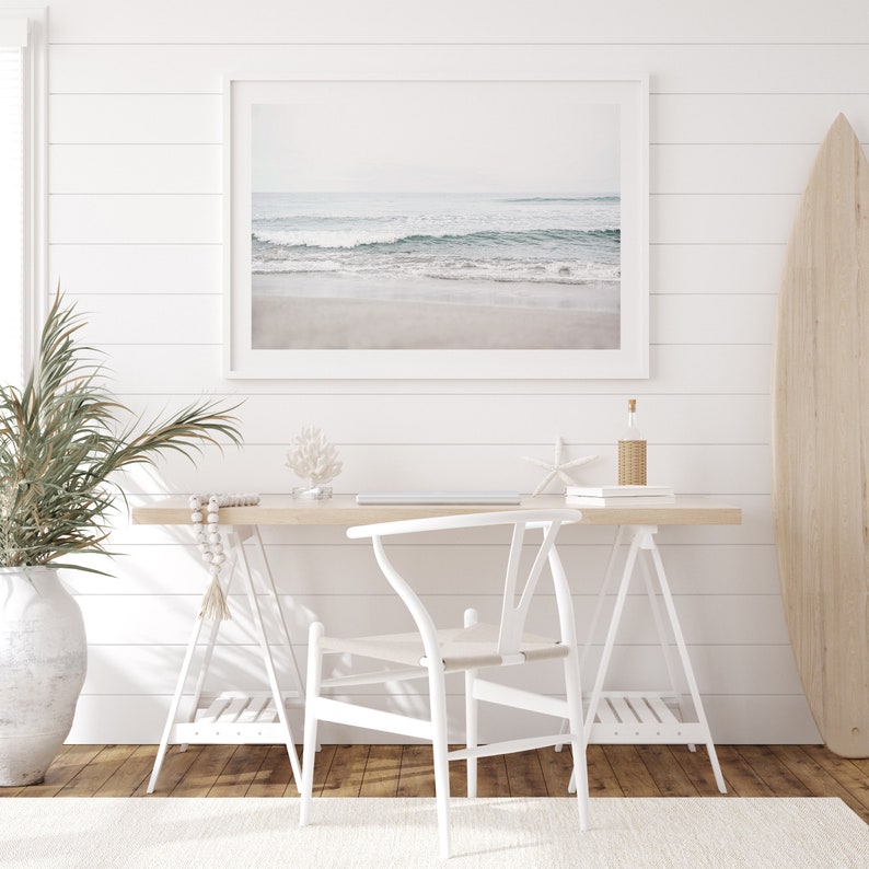 May include: A white desk with a white chair and a framed photograph of a beach scene. There is a surfboard leaning against the wall and a potted plant on the floor.