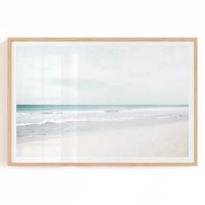 May include: A framed print depicting a tranquil beach vista. The artwork shows a white sandy shore, a turquoise sea, and a pale blue sky. The image is enclosed in a light brown wooden frame, offering a calming coastal feel.