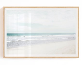 May include: A framed print depicting a tranquil beach vista. The artwork shows a white sandy shore, a turquoise sea, and a pale blue sky. The image is enclosed in a light brown wooden frame, offering a calming coastal feel.