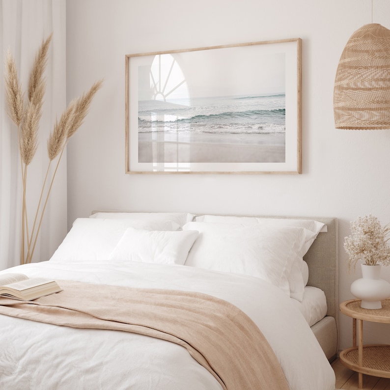 May include: A white bed with a beige throw blanket and white pillows. A framed photograph of a beach scene hangs above the bed. A wicker lamp hangs from the ceiling.