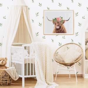 May include: A white crib with a white canopy and a white blanket. A brown teddy bear sits in a wicker basket next to the crib. A wicker chair with a white cushion sits in the corner of the room. A framed print of a Highland cow with a flower crown hangs on the wall. The wall is decorated with green leaf decals.