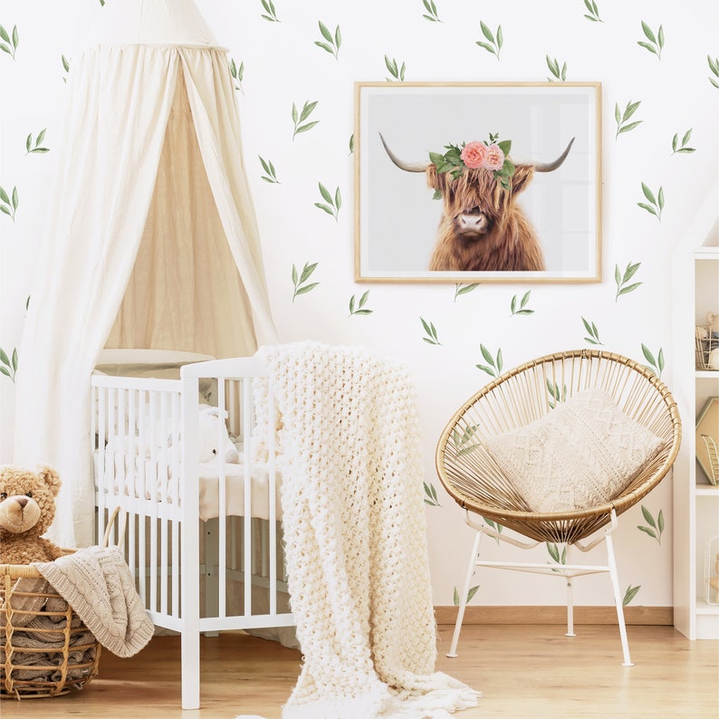 May include: A white crib with a white canopy and a white blanket. A brown teddy bear sits in a wicker basket next to the crib. A wicker chair with a white cushion sits in the corner of the room. A framed print of a Highland cow with a flower crown hangs on the wall. The wall is decorated with green leaf decals.