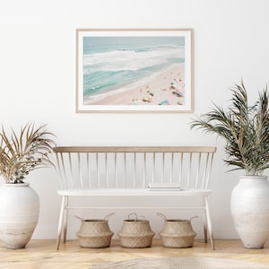 May include: A framed photograph of a beach scene with people sunbathing and colorful umbrellas. The photo is hanging on a white wall above a white bench with three woven baskets underneath.