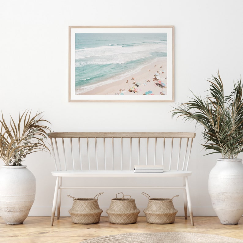 May include: A framed photograph of a beach scene with people sunbathing and colorful umbrellas. The photo is hanging on a white wall above a white bench with three woven baskets underneath.
