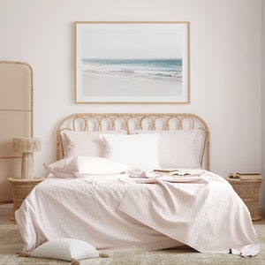 May include: A light pink linen bedspread with a white pillow and a framed photograph of a beach scene above the bed.