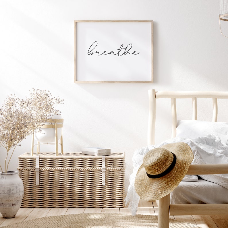 May include: A framed print with the word "breathe" in a cursive font hangs above a bed. A woven trunk, a straw hat, and a vase with dried flowers are in the room. The room has a neutral color palette and a relaxed, natural aesthetic.