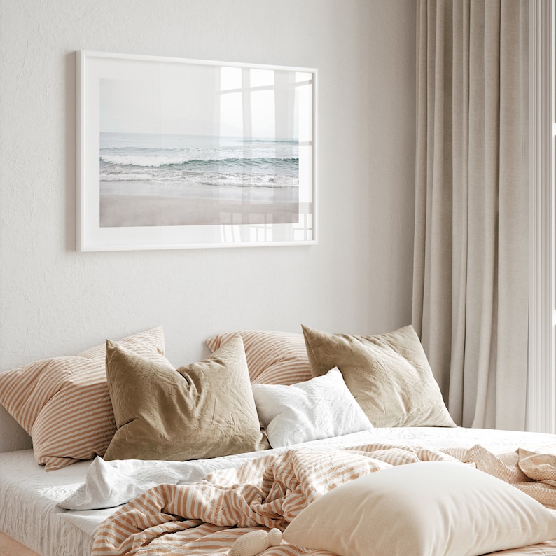 May include: A bedroom with a white framed photograph of a beach scene above a bed with a white, brown, and beige striped comforter and pillows. The bed is against a white wall and there are beige curtains in the background.