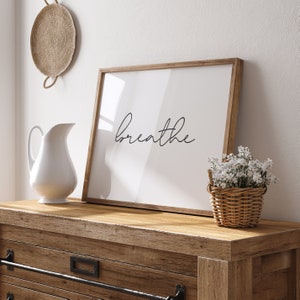 May include: A framed print with the word "breathe" in cursive script sits on a wooden dresser. A white pitcher and a small basket of white flowers are also on the dresser. A woven wall hanging is visible in the background.