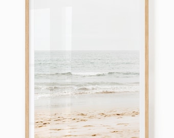 May include: A framed print depicting a beach scene. The image shows a sandy beach with gentle waves from the sea. The sky is a soft, muted colour, creating a calm atmosphere. The frame is made of light-coloured wood, adding to the tranquil aesthetic.