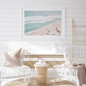 May include: A framed photograph of a beach scene with turquoise water, white sand, and colorful umbrellas. Two white wicker chairs with pink pillows are positioned in front of the photo, along with a small wicker table and a white shelf.