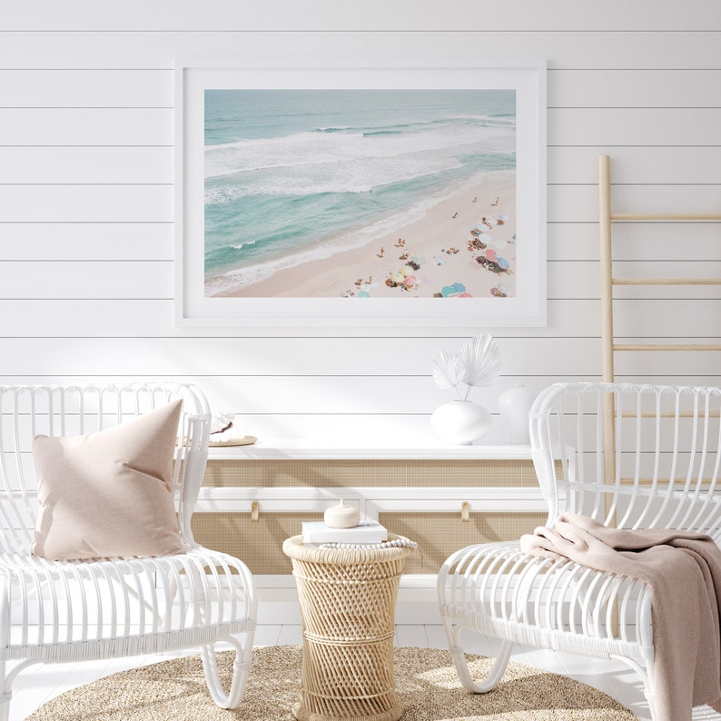May include: A framed photograph of a beach scene with turquoise water, white sand, and colorful umbrellas. Two white wicker chairs with pink pillows are positioned in front of the photo, along with a small wicker table and a white shelf.
