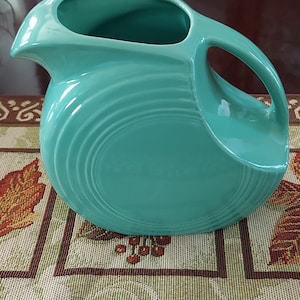May include: A turquoise ceramic pitcher with a wide mouth and a handle. The pitcher has a smooth, glossy finish and a rounded shape.