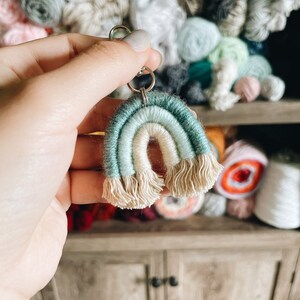 May include: A teal and white crocheted rainbow keychain with fringe. The keychain is held in a hand against a background of yarn balls.