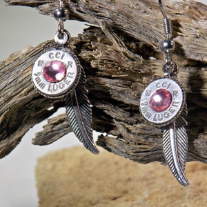 Può includere: Un paio di orecchini d'argento con un strass rosa incastonato in un bossolo di proiettile Luger da 9 mm con un ciondolo a forma di piuma che pende sotto.