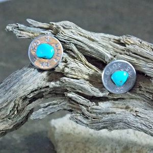 May include: Two turquoise stones set in silver bullet casings. The casings have markings on them, including the year "1974" and "1970".