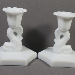 May include: A pair of white glass candle holders. Each features a square base, a dolphin-shaped stem, and a tulip-shaped cup. The candle holders are made of opaque white glass and are set against a grey background.