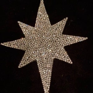Puede incluir: Una gran estrella brillante en forma de estrella plateada hecha de diamantes de imitación. La estrella está sobre un fondo negro.