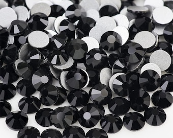 May include: A large quantity of black rhinestones, also known as crystals or gems, are scattered on a white surface. The rhinestones are round and have a faceted design.