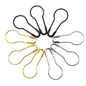 May include: A selection of pear-shaped safety pins in black, gold, and silver. The pins are arranged in a circular pattern. The pins are made of metal and have a simple, functional design. The pin heads form a ring.