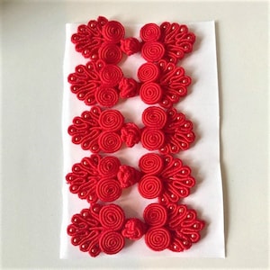 Könnte beinhalten: Sechs rote chinesische Knotenknauf mit weißen Perlen. Die Knoten haben eine Blütenform mit einem Wirbelmuster.