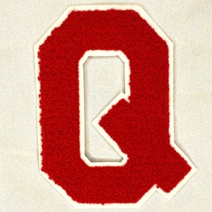 May include: A red chenille letter "Q" patch with a white border, set against a light beige fabric background. The letter has a textured, fuzzy appearance, typical of varsity letter patches.
