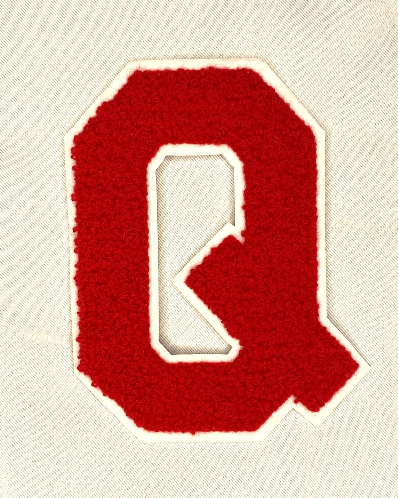 May include: A red chenille letter "Q" patch with a white border, set against a light beige fabric background. The letter has a textured, fuzzy appearance, typical of varsity letter patches.