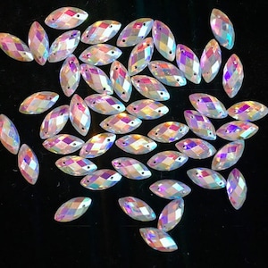 May include: A large quantity of iridescent, clear, faceted, marquise-shaped rhinestones. The rhinestones are scattered on a black background.