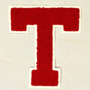 May include: A red felt letter "T" patch with a white border, set against a light gray fabric background. The letter has a textured, fuzzy appearance, typical of varsity letter patches. Ideal for jackets or bags.